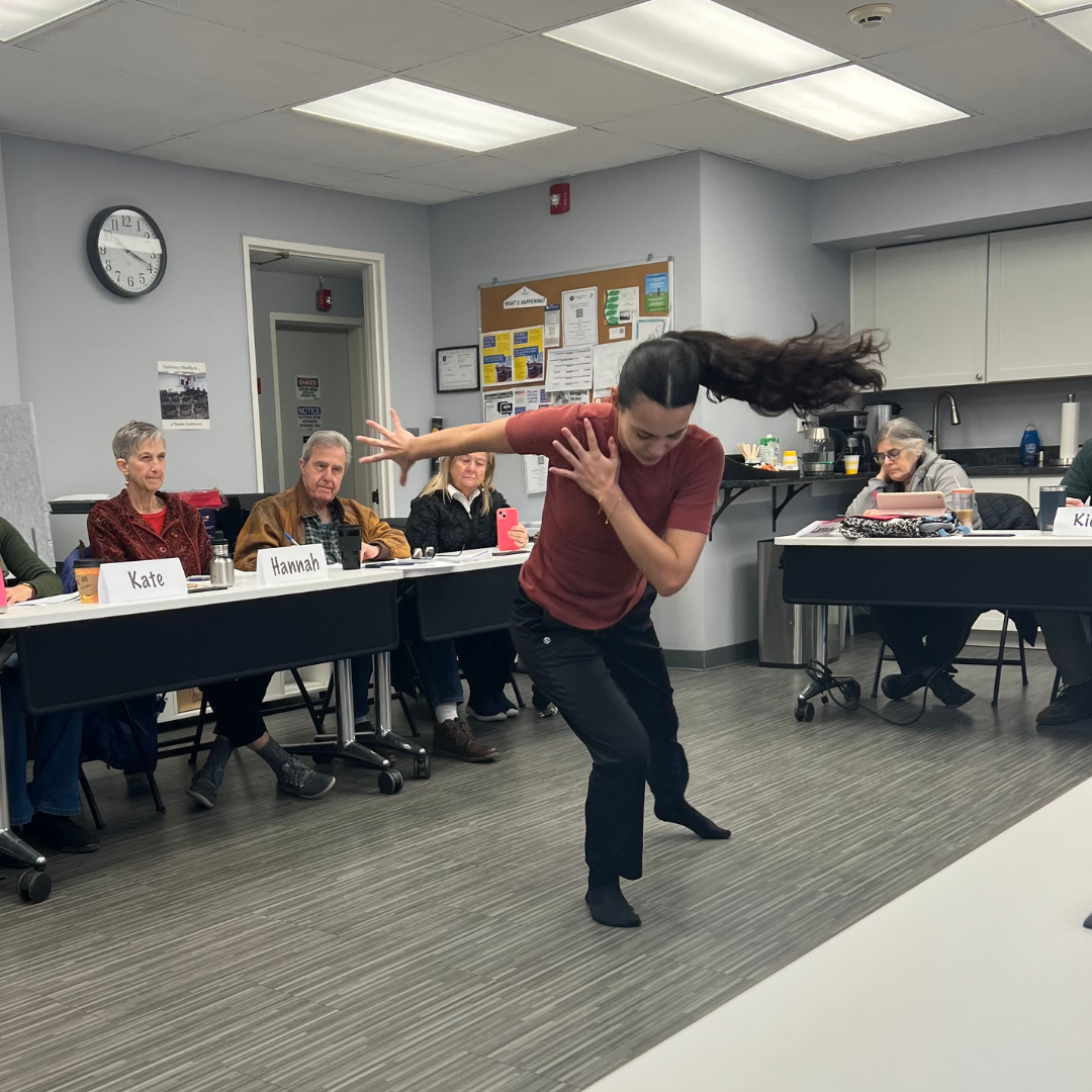 Woman in black pants and burnt orange shirt dances as 4 adult students look on.