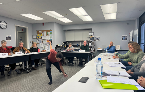 Woman in black pants and burnt orange shirt leans forward with arms outstretched as she dances in front of a group of adults seated at tables surrounding her.